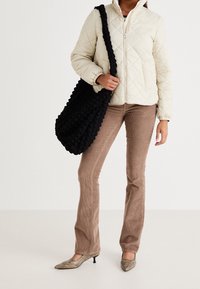 Beige quiltet jakke med høj krave, kombineret med brune fløjlsbukser med vidde og en tekstureret sort skuldertaske. Spidse guld sko.