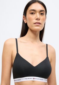 Fekete bralette vékony pántokkal, V-nyakú kialakítással és az alsó részen fehér gumipánttal, amelyen a "TOMMY HILFIGER" logó található.