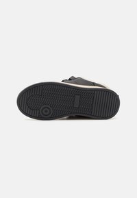 Svart såle til sneakers med teksturert gummi, med fiskebeinmønster og sirkulært emblem. Forsterket hæl med beige aksent nær den øvre delen.