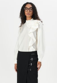 Hvit bluse med volanger ved skuldrene, kombinert med et svart skjørt med flere sølvspenner og en sidetrykkdesign.