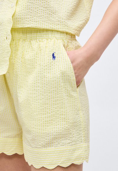 Χέρι στην τσέπη κίτρινων σορτς σερσάκερ με κυματιστό τελείωμα και μπλε κεντημένο λογότυπο, συνδυασμένα με αντίστοιχο υφασμάτινο πουκάμισο.