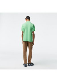 Grön polo skjorta i kombination med bruna mönstrade byxor med geometrisk design. Modell som bär svarta sneakers, sedd bakifrån.