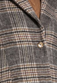Šedý kostkovaný vlněný kabát s klopovým límcem, zlatým zapínáním na knoflíky a kontrastními hnědými a krémovými pruhy po celé délce.