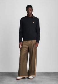 Černá mikina s hnědým límcem a malým logem zvířete na hrudi, doplněná širokými béžovými kalhotami s postranními pruhy a bílými botami.