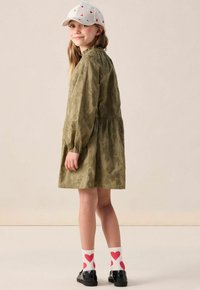 Kind, der ein olivgrünes Blumenkleid trägt, weiße Socken mit roten Herzen, schwarze Schuhe und eine weiße Mütze mit kleinen roten Herzen, und seitlich lächelt.