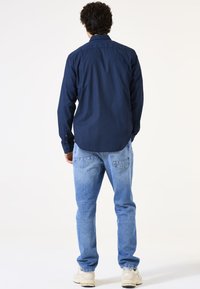 Dunkelblaue, gemusterte Langarmhemd mit Knöpfen, kombiniert mit hellblauen Jeans und beigen Sneakers, von hinten betrachtet.