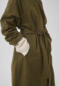 Olijfgroene jas met ceintuur en zijzakken, gedragen over wit geribd gebreide mouwen, getoond van middel tot bovenbenen.