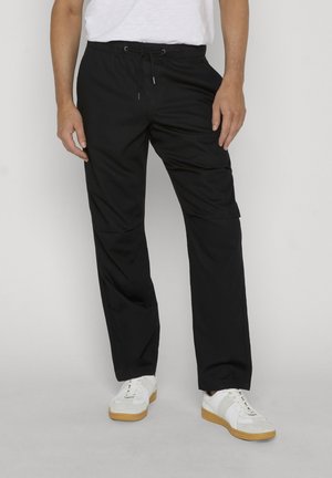 Homme portant un pantalon cargo à cordon noir et des baskets blanches, debout devant un fond clair uni.