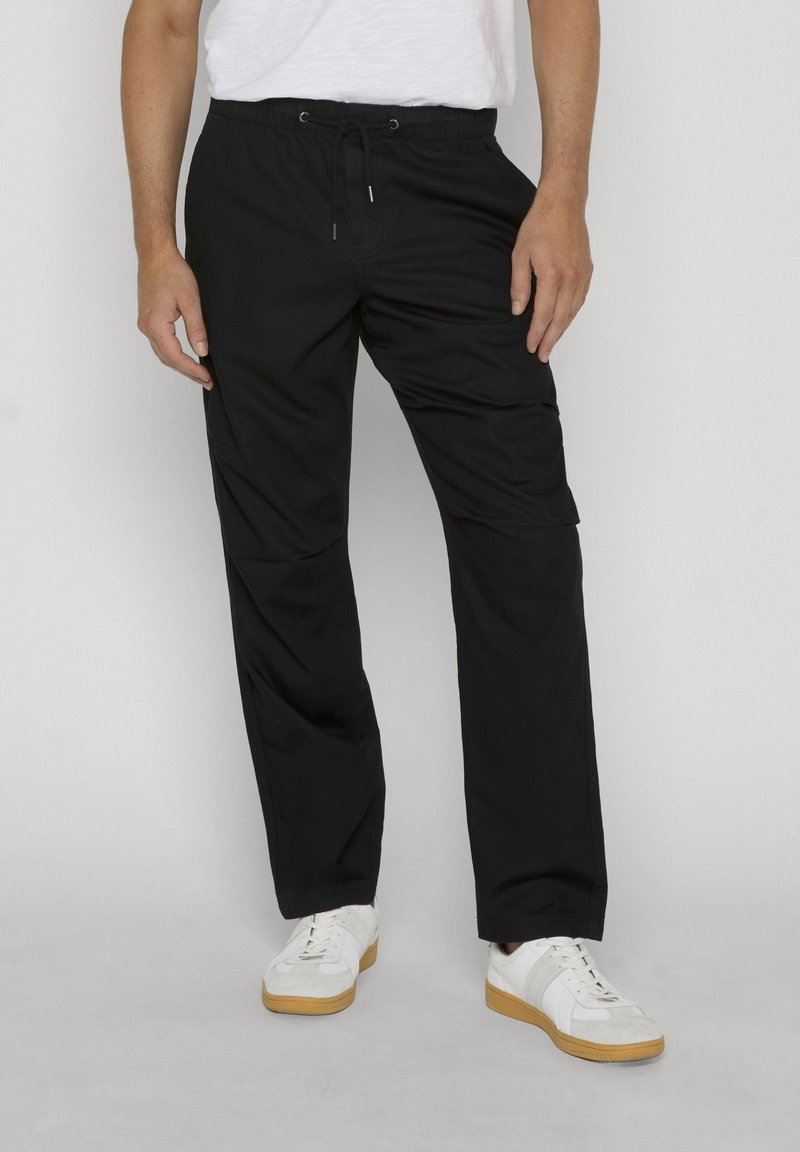 Homme portant un pantalon cargo à cordon noir et des baskets blanches, debout devant un fond clair uni.