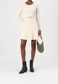 Robe en maille crème côtelée avec manches longues, taille cintrée, jupe évasée et ceinture accentuée d'or. Associée à des bottines noires et un sac olive.