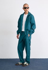 Svetlo modra trenirka iz lahkega blaga, ki vključuje jakno z zadrgo in hlače z elastičnim pasom. Črni supergi z belimi črtami dopolnjujejo videz.