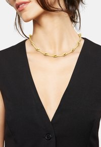 Guld kæde med aflange ovale led, båret direkte på bar hud imod en sort dyb V-hals outfit.
