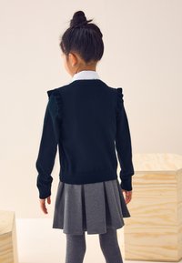 Ciemnozielony dzianinowy sweter z falbanami na rękawach, w połączeniu z szarą plisowaną spódnicą i szarymi legginsami. Widok od tyłu.