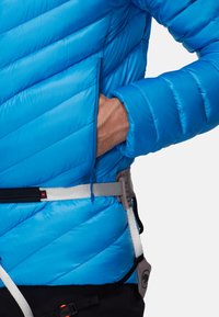 Veste isolante bleue avec motifs matelassés diagonaux, poche zippée et ceinture ajustable avec détails métalliques.