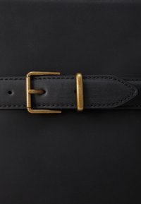 Ceinture en cuir noir avec une boucle en laiton, présentant une texture lisse, des coutures décoratives et une extrémité pointue. Ajustable avec plusieurs trous.