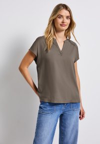 Top s krátkým rukávem a véčkovým výstřihem v olivově zelené barvě, hladký materiál, volný střih. Spárován se světle modrými kalhotami s širokými nohavicemi.