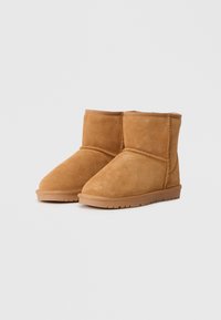 Botas de tornozelo de camurça em tom claro, com um design simples, bico arredondado e detalhes costurados, apresentando uma sola de borracha plana.
