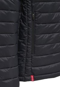 Sort quiltet jakkeafsnit med lodret lynlåslomme og rød mærkeetiket i bunden på en glat, polstret stof.