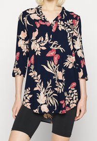 Donna che indossa una tunica blu navy con stampe floreali rosa e farfalle rosse, maniche tre quarti e pantaloni ciclistici neri attillati.