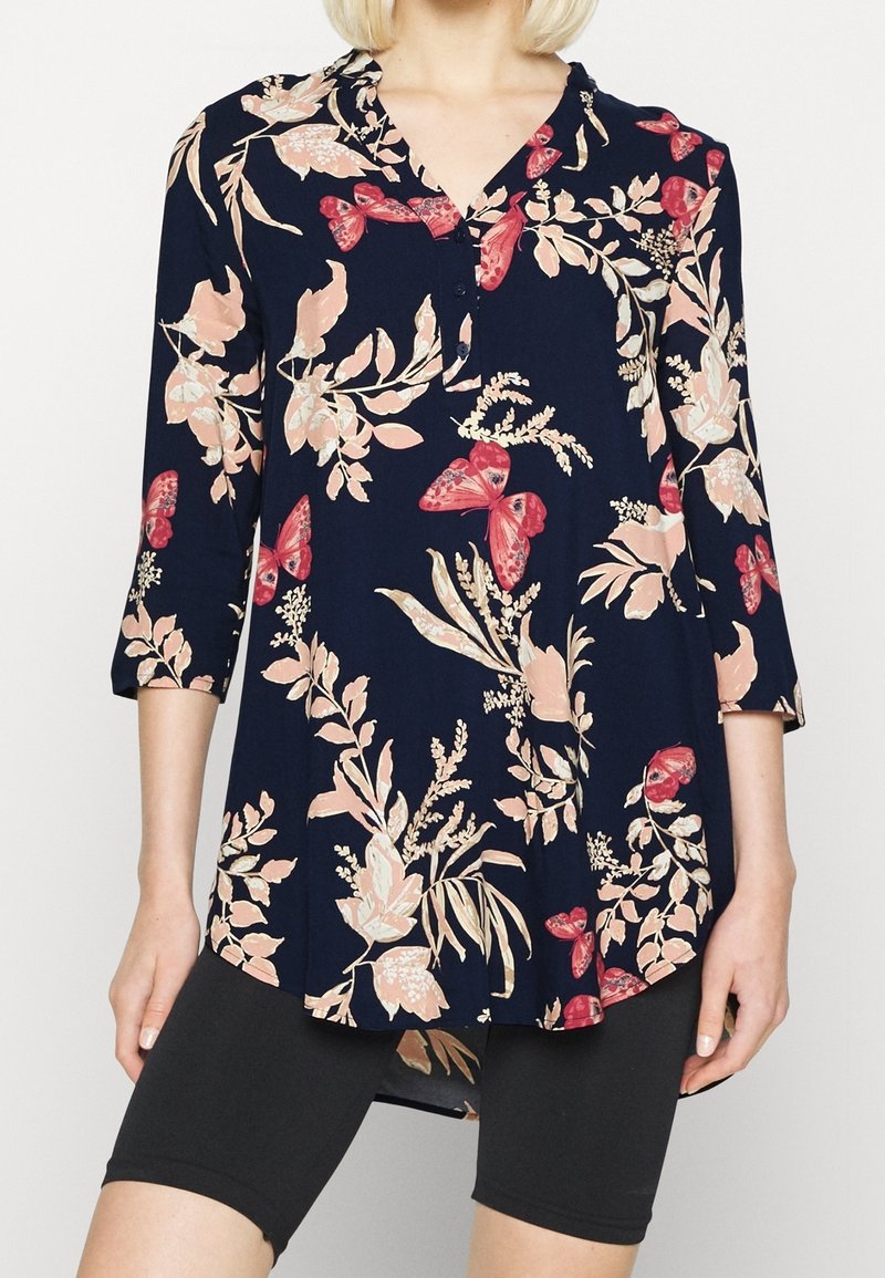 Donna che indossa una tunica blu navy con stampe floreali rosa e farfalle rosse, maniche tre quarti e pantaloni ciclistici neri attillati.