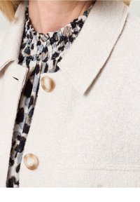 Weißer strukturierter Blazer mit Kragen und einem großen beigefarbenen Knopf, über einem Rüschenshirt mit dunklem Blumenmuster getragen.