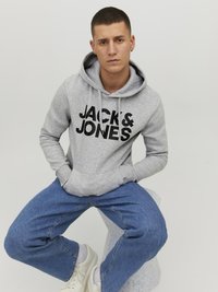 Gray hoodie with "JACK & JONES" logo, kangaroo pocket, and drawstring hood. Worn with blue jeans and white sneakers, seated on a stool.