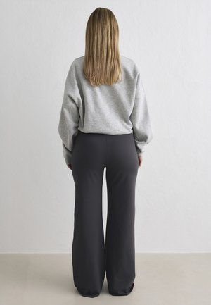 Žena s dlouhými blond vlasy, která má na sobě šedou mikinu a široké černé kalhoty, stojí čelem k jednobarevné bílé stěně.