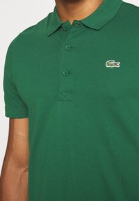 Man med en grön kortärmad pikétröja med krage och tre knappar, med en liten krokodil-logotyp på vänster bröst.