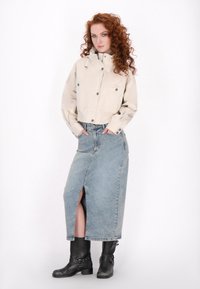 Jachetă bej scurtă cu două buzunare frontale, asortată cu o fustă maxi din denim albastru cu o fanta laterală și cizme negre până la genunchi.