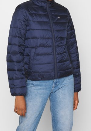 Personne portant une veste matelassée bleu marine zippée, associée à un jean bleu clair, montrée des épaules aux hanches.