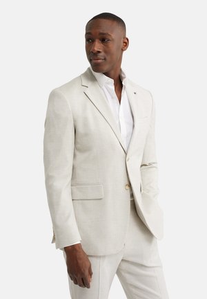 Man draagt een licht beige pak en wit overhemd, staat met één hand in de zak en kijkt iets naar links.