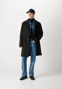 Manteau long noir avec un design croisé, superposé à une veste en denim et un col roulé noir, associé à un jean bleu clair et des chaussures noires.