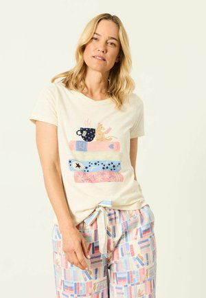 Blonde woman wearing a beige t-shirt with a mouse, books, and mug print, and pastel-colored pajama pants with a book pattern.