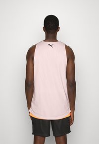 Hombre vestido con una camiseta deportiva sin mangas de Puma en un suave tono rosa y pantalones cortos de atletismo negros, de pie frente a una pared blanca sin adornos.