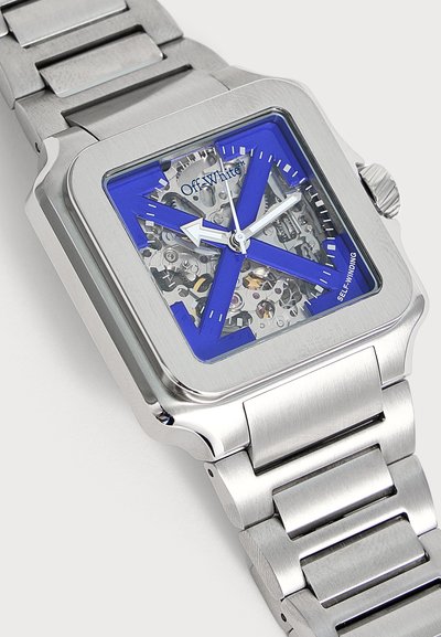 Montre-bracelet carrée argentée avec un design géométrique bleu, des engrenages mécaniques visibles, des aiguilles blanches pour les heures et les minutes, et un bracelet en acier inoxydable.