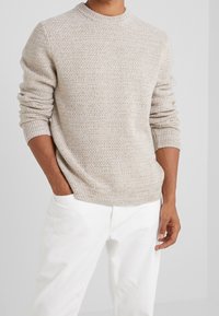 Beiger Strickpullover mit Rundhalsausschnitt, der ein strukturiertes Muster und gerippte Ärmelbündchen aufweist, kombiniert mit weißen Hosen.