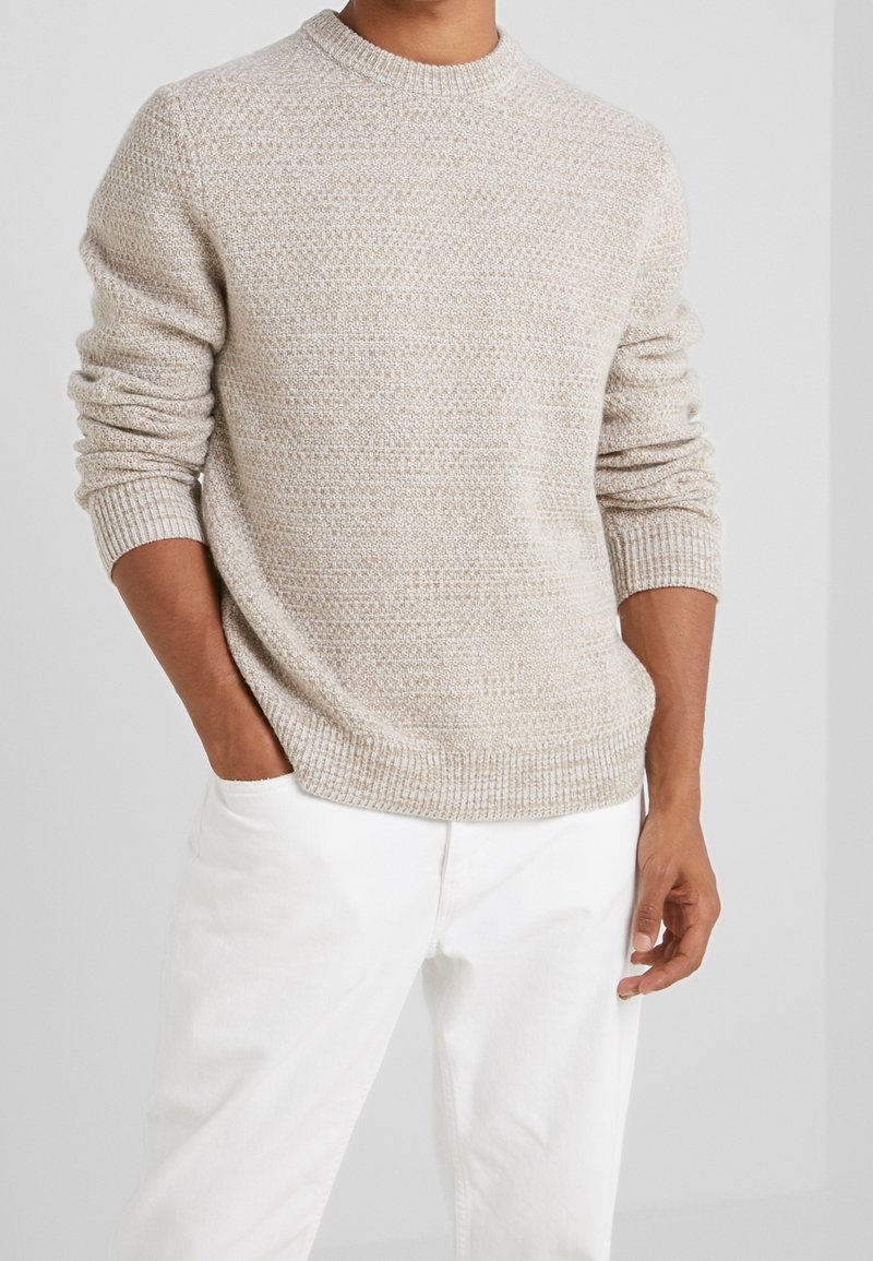 Beiger Strickpullover mit Rundhalsausschnitt, der ein strukturiertes Muster und gerippte Ärmelbündchen aufweist, kombiniert mit weißen Hosen.