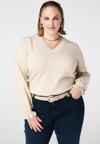 Beige V-hals trui met pofmouwen en geplooide manchetten, gecombineerd met donkerblauwe spijkerbroek en een beige riem met een gestrikte accent.