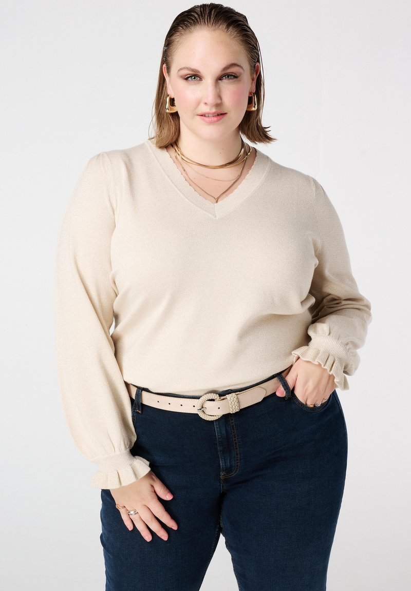 Beige V-hals trui met pofmouwen en geplooide manchetten, gecombineerd met donkerblauwe spijkerbroek en een beige riem met een gestrikte accent.