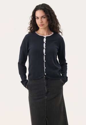 Navy cardigan with a round neckline, white trim, and decorative buttons down the front. Soft knit material, worn over a dark denim skirt.