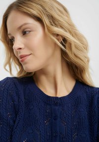 Cardigan lavorato a maglia navy con un motivo testurizzato, scollatura rotonda e chiusura con bottoni. Presenta dettagli intricati simili a pizzi sul tessuto.