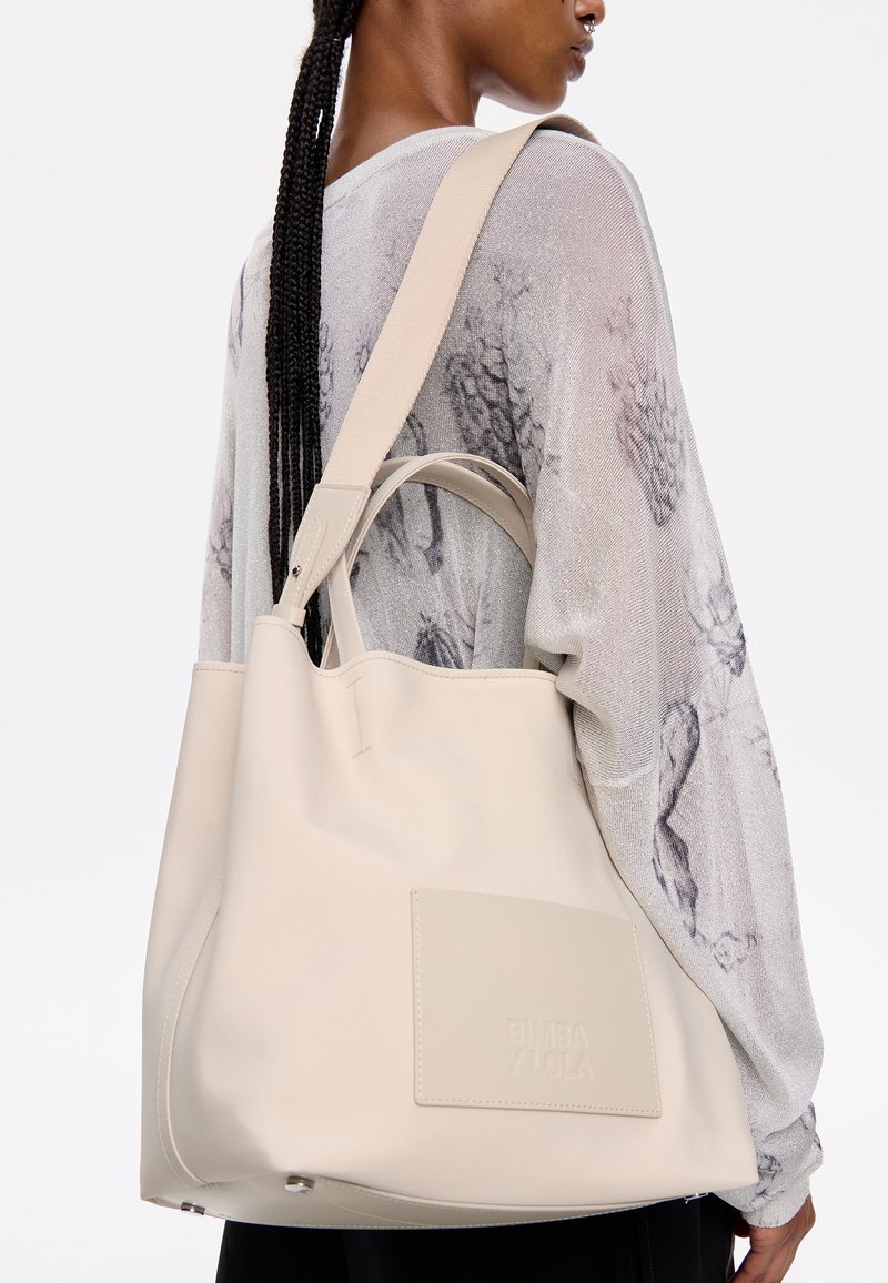 Bolso tote beige hecho de material suave, con doble asa, un parche con el logo y una correa para el hombro. El diseño es minimalista y moderno.