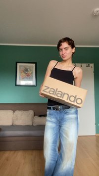 Zalando-kartonki, som fargen lys brun med en fet, mørk grå logo. En person i en tettsittende svart topp og lyseblå denimjeans holder boksen.