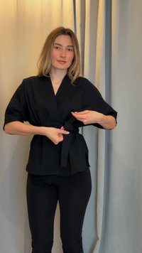 Γυναίκα με ανοιχτό καστανά μαλλιά δένει μια μαύρη ζώνη γύρω από ένα μαύρο τοπ στυλ κιμονό, στέκεται μπροστά από μπεζ και γκρι κουρτίνες.