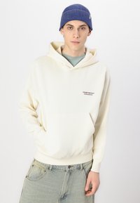 Sudadera color crema con bolsillo canguro, capucha con cordón y logo bordado. Combinada con jeans de color azul claro. Llevada con un gorro de punto azul.