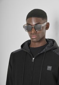Felpa nera con zip, caratterizzata da una superficie texturizzata, con un cappuccio nero liscio e accenti sulle spalle. Abbinata a occhiali da sole trasparenti oversize.