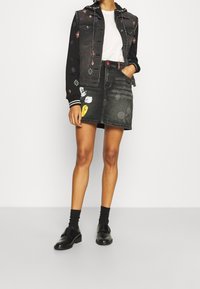 Persona che indossa una giacca di jeans nera con ricami colorati, una camicia bianca, una gonna di jeans nera con toppe di cartoni, calze nere e scarpe nere.