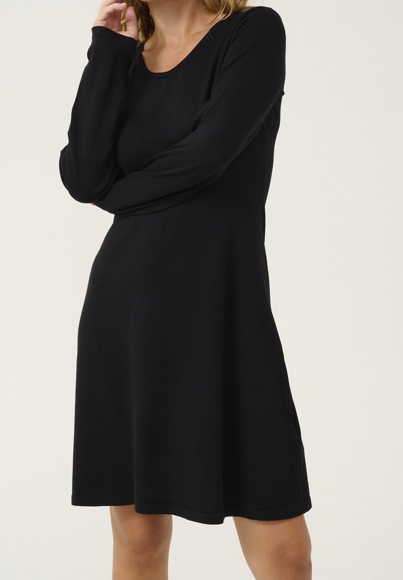 Robe noire à manches longues en tissu doux, avec un col rond et une silhouette trapèze, présentant une texture lisse et des coutures minimalistes.