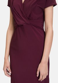 Bordeaux wikkeljurk met korte mouwen, voorzien van een geplooid taille-detail en glad, elastisch materiaal, wat zorgt voor een getailleerde silhuet.
