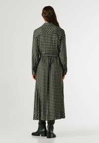 Καρό μακρύ φόρεμα σε μαύρο και κρεμ με σφιχτό τμήμα στη μέση, κουμπωτές μανσέτες και γιακά, συνδυασμένο με μαύρες ψηλές μπότες.